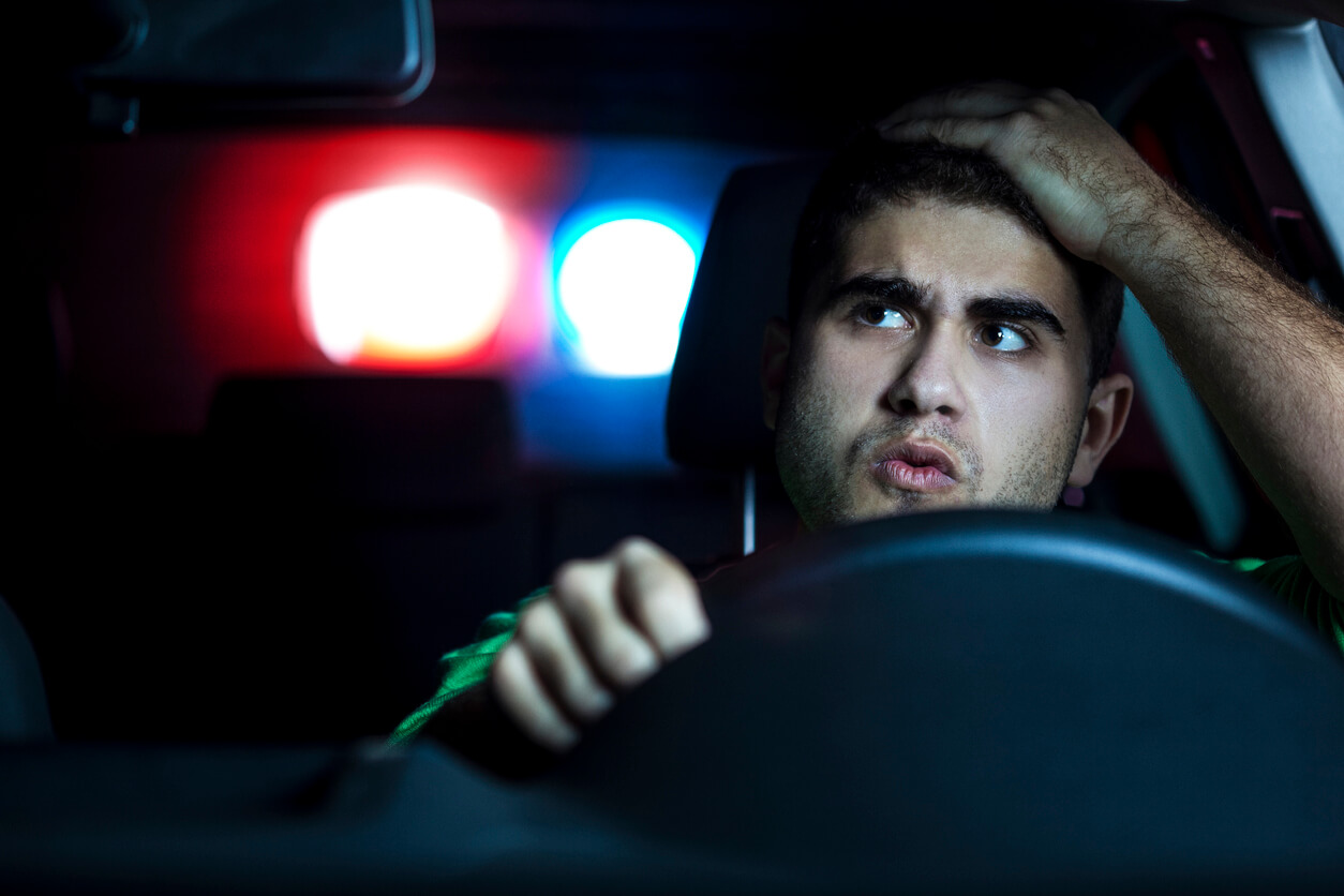 Man looking stressed in the divers seat of his car with his hand on his forehead and cop car lights behind him